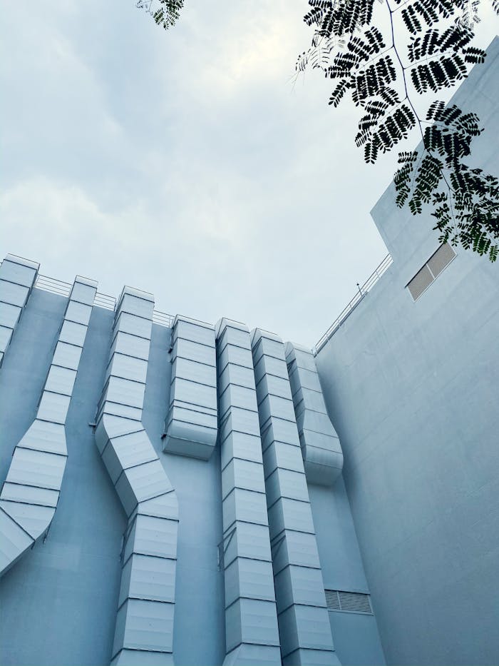 Low-angle view of industrial ductwork on a modern building exterior with sky and tree leaves.